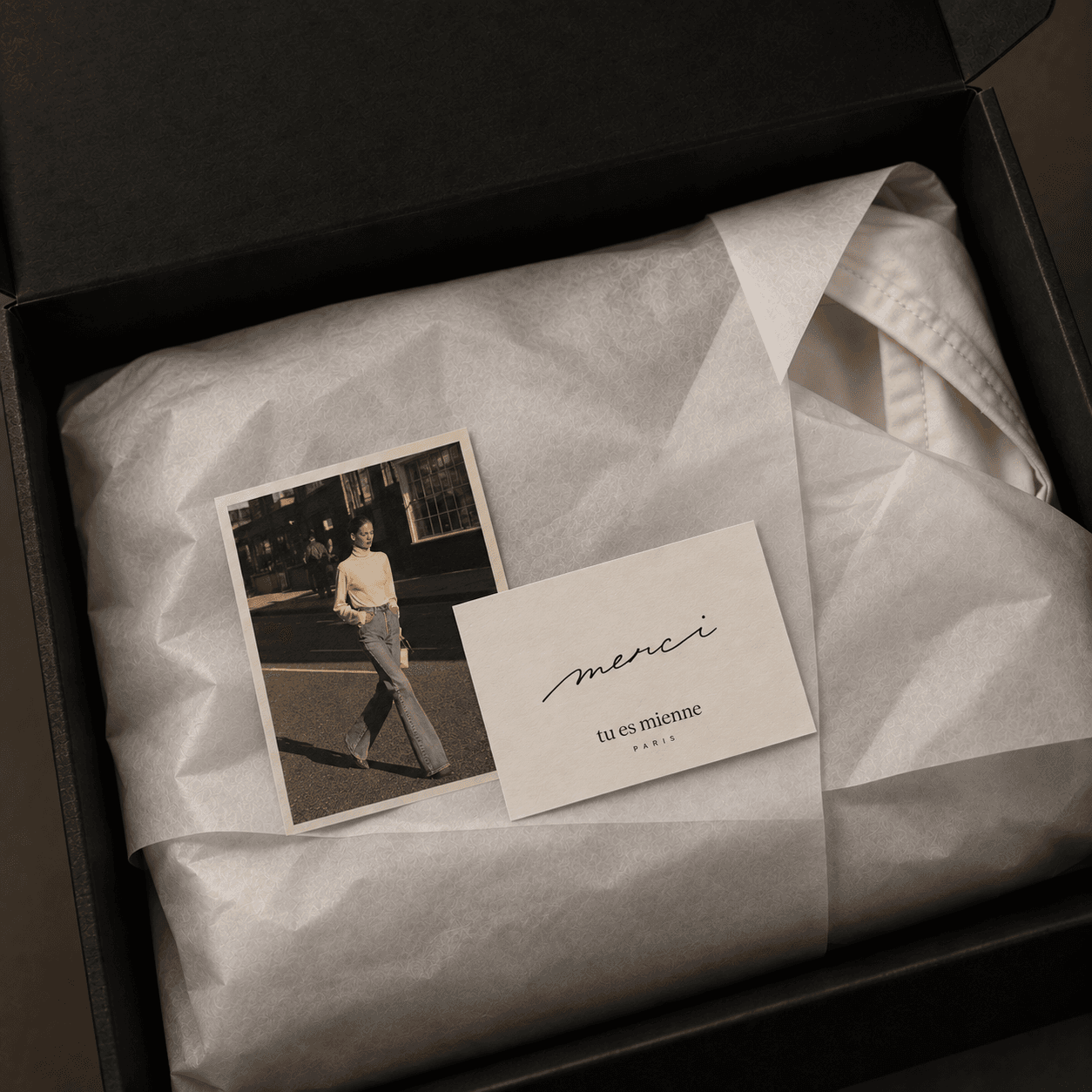 An open black gift box lined with tissue paper, a polaroid of the campaign and a card reading merci, tu es mienne Paris.