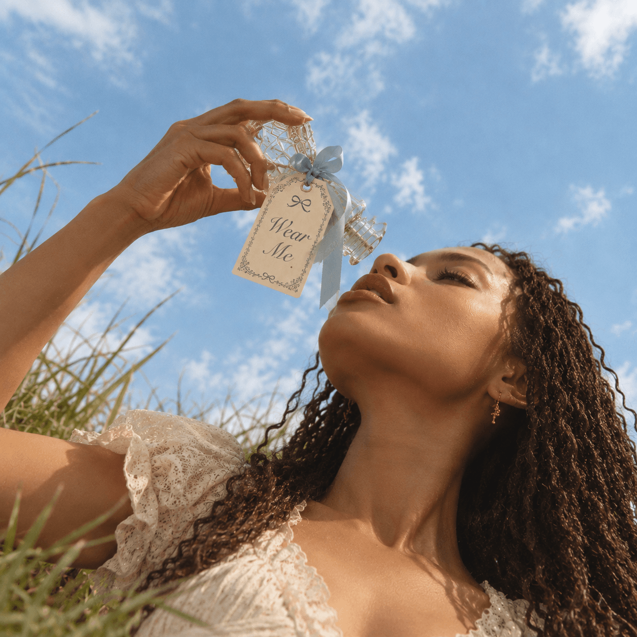 A woman lying in long grass holds a cut-glass perfume bottle above her face against blue sky, a pale blue ribbon tying on a tag that reads Wear Me.