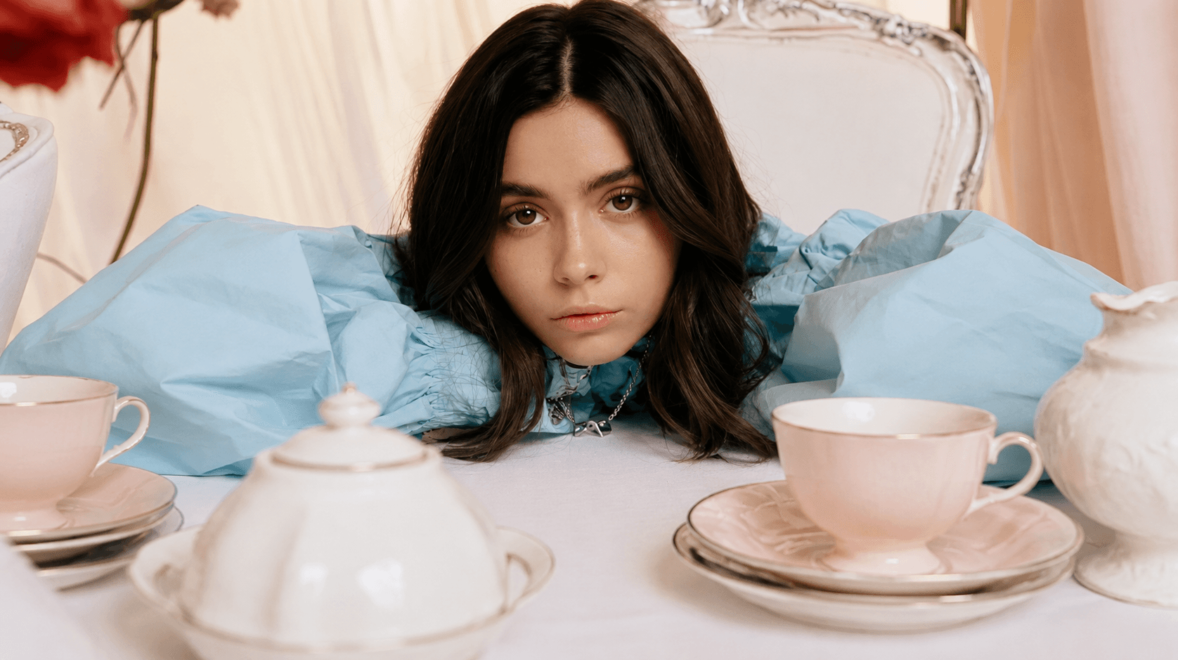 A woman rests her chin on a white tablecloth between pink teacups and a white sugar bowl, her pale blue puffed sleeves spread across the table.