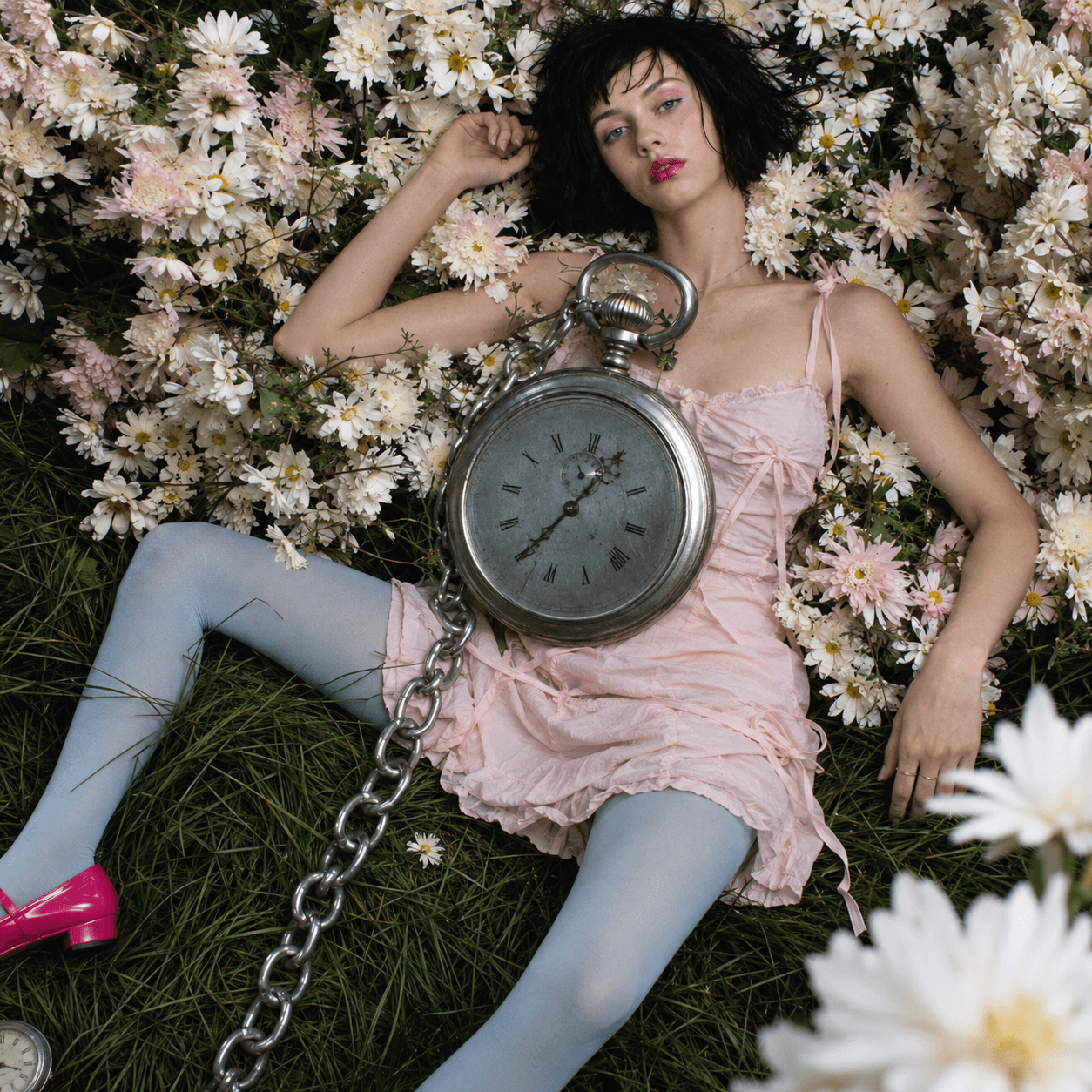 A woman in a pink ribbon-tied slip dress and pale blue tights lies on grass against a bank of daisies, a giant pocket watch on a heavy chain resting across her.