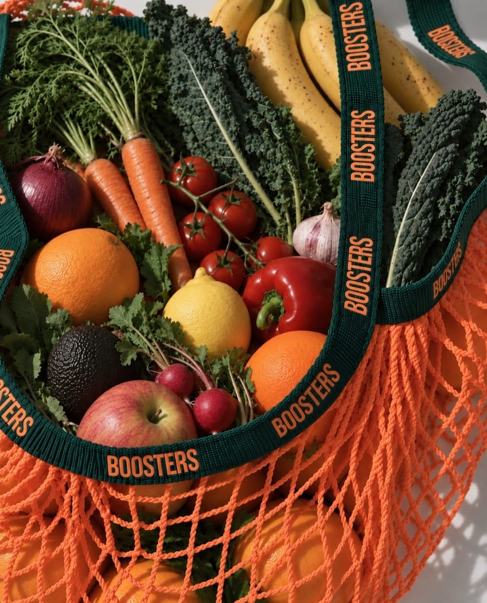 An orange string bag printed with the Boosters wordmark, packed with kale, tomatoes, citrus, carrots and bananas.