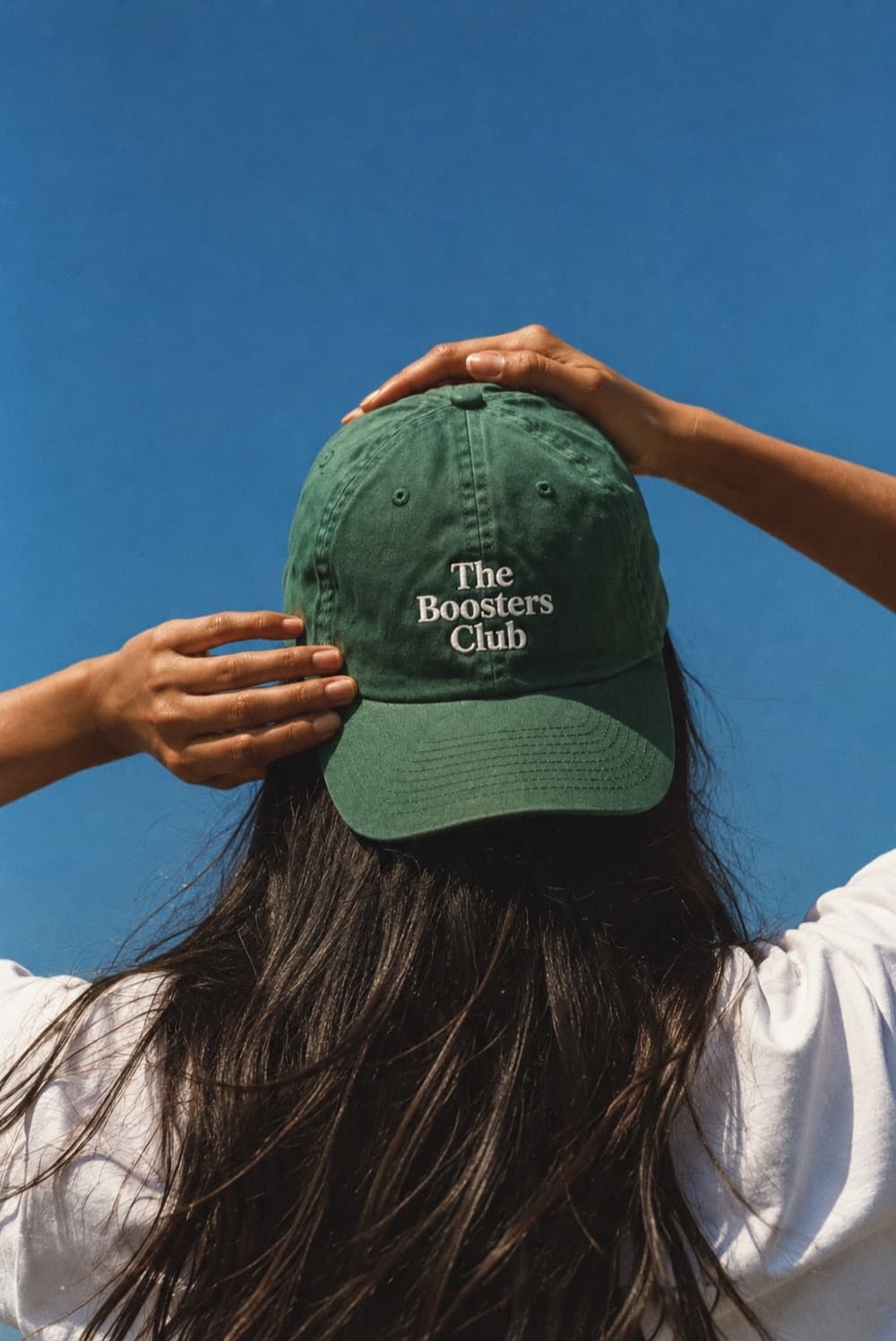Seen from behind against a clear blue sky, a woman has both hands on a dark green cap, worn backwards and embroidered with The Boosters Club in white.