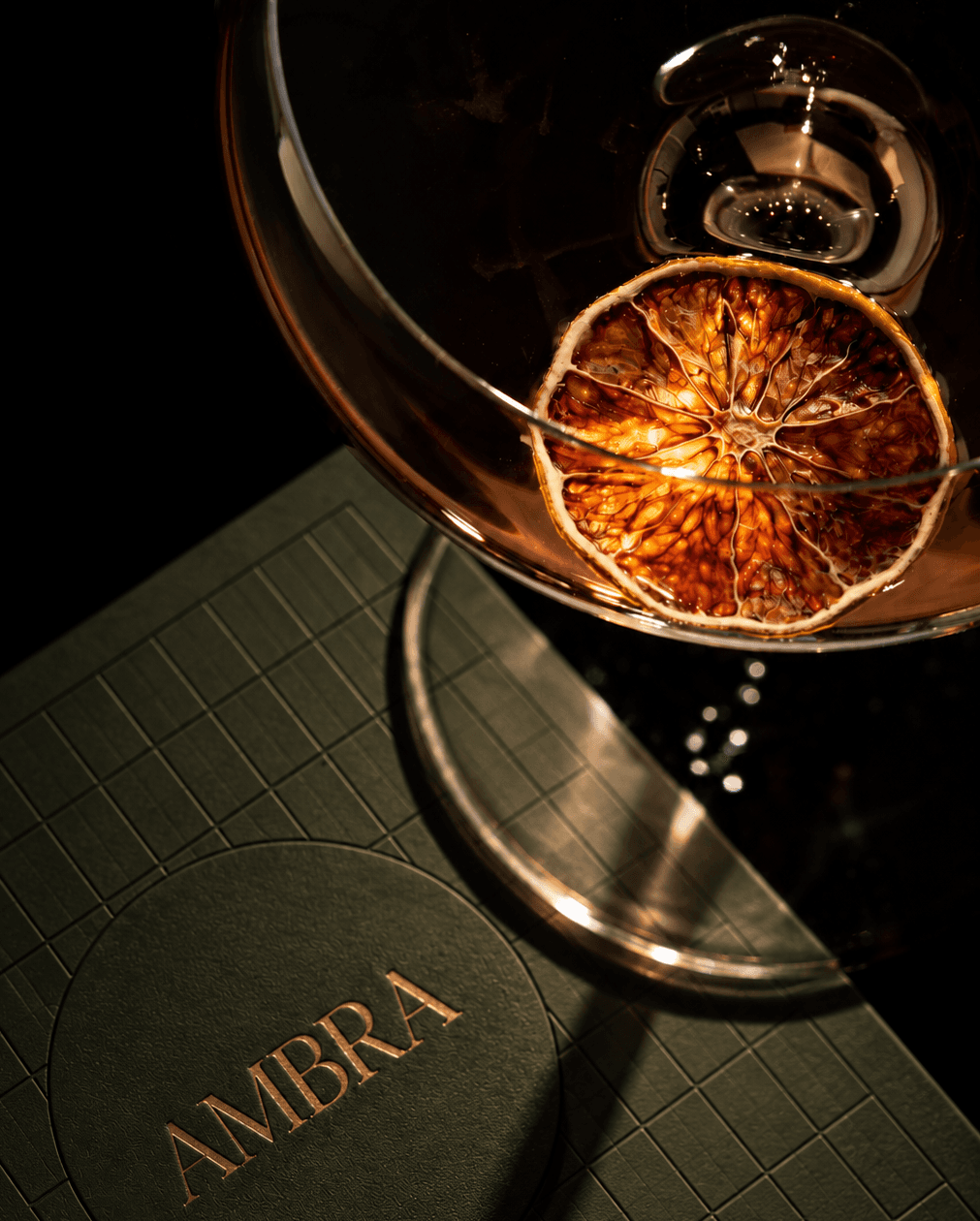A coupe with a dried orange slice seen from above, its glass and shadow falling across the olive menu cover, the AMBRA wordmark foiled in gold.