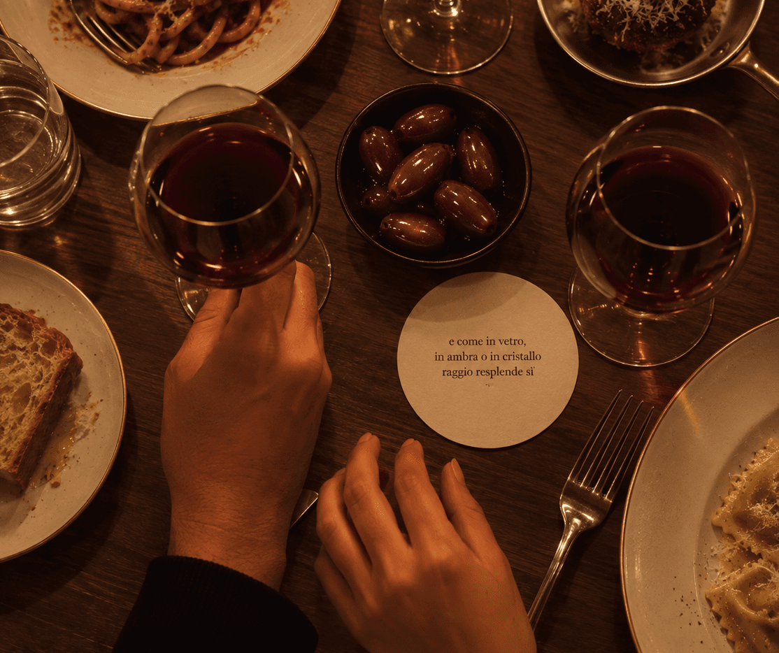 An overhead shot of Ambra's table set with wine, olives and pasta, a round coaster printed with a line of Italian verse at its centre.