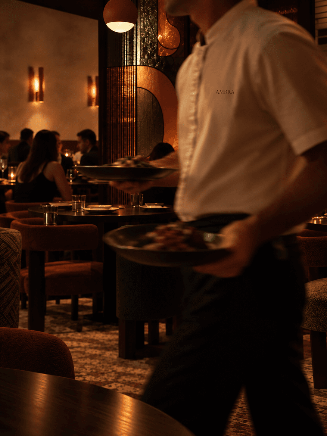 A waiter crosses Ambra's dining room carrying two plates, the wordmark embroidered on his shirt, the stained-glass screen glowing behind him.